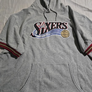 May include: A gray hoodie with the word "SIXERS" in a stylized design across the chest. The design includes a star, a basketball, and blue and white stripes. The sleeves have red and gold stripes.