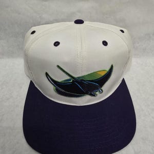 May include: A white baseball cap with a navy blue brim. The cap features a stylized black and green stingray logo on the front. The top of the cap has navy blue button and stitching details.
