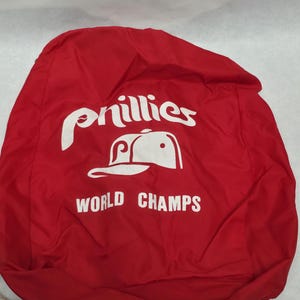 May include: Red nylon backpack with white text and a baseball cap graphic. The text reads "Phillies" above a baseball cap graphic and "World Champs" below. The backpack has a white strap.
