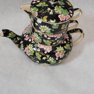 May include: A black teapot with a floral pattern in pink, green, and yellow. The teapot has a lid and is stacked on top of two matching cups.