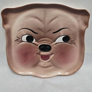 May include: A vintage ceramic plate in the shape of a cartoon pig face. The plate features a light brown color with rosy cheeks, black eyes, and a black nose. The pig has a slightly grumpy expression.