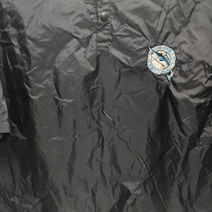 May include: Black baseball jacket with a button-up front and a small logo on the right chest. The logo is a circular patch with a marlin fish design in blue, white, and teal. The jacket has a drawstring at the bottom.