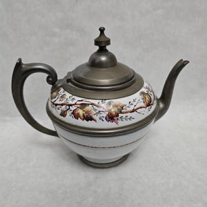 May include: Antique white porcelain teapot with a metal lid, handle, and spout. The teapot features a floral design with brown, yellow, and pink leaves. The lid has a small knob on top.