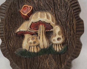 May include: A brown wooden plaque with a raised design of three mushrooms. The mushrooms are white with red accents and are set against a green background. The plaque has a scalloped edge.
