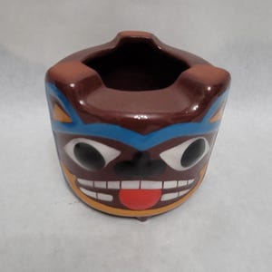 May include: Brown ceramic ashtray with a cartoon face. The face has blue eyes, a red tongue, and a yellow mouth. The ashtray is decorated with blue and yellow lines.