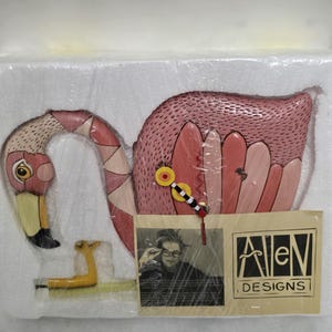 May include: A pink flamingo-shaped wall clock with a yellow beak and legs. The clock has a patterned neck and wing, with a yellow and black clock face. The Allen Designs logo is visible.