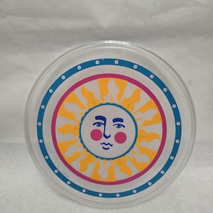 May include: A circular clear plastic plate featuring a central sun illustration with a face, yellow rays, and pink cheeks. The design is framed by a vibrant pink ring and an outer blue border decorated with small white dots and tiny pink circles.