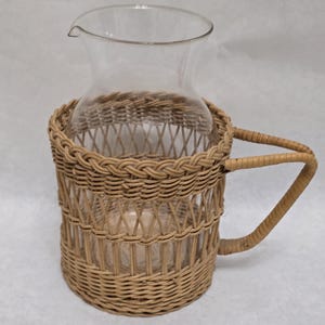 May include: A clear glass pitcher with a spout, encased in a woven wicker basket. The basket has a handle and a natural tan color. The pitcher is designed for serving beverages.
