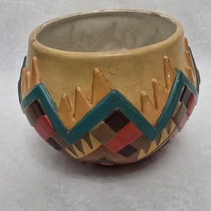 May include: A round ceramic planter with a wide opening and a smooth, light tan upper rim. The lower section features a raised, textured geometric pattern in teal, brown, and red, with triangular accents pointing upward toward the rim.