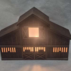 May include: A dark brown wooden barn-shaped decorative light fixture featuring a gabled roof, a central square window, and horizontal slatted openings along the lower level. Warm light glows from within the structure, illuminating the cutouts.