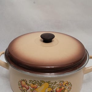 May include: A vintage enamelware pot with a brown lid and a mushroom design. The pot has a cream-colored base with a brown rim and a silver trim. The mushroom design features yellow, orange, and brown mushrooms with green leaves.