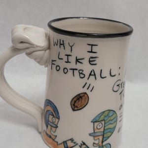 May include: White ceramic mug with a hand-shaped handle. The mug has a black rim and a hand-drawn illustration of two football players and a football. The text "WHY I LIKE FOOTBALL!!!" is written on the mug.