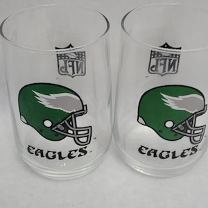 May include: Two clear glass tumblers featuring the Philadelphia Eagles football team logo. Each glass has a green helmet design with the word "EAGLES" in a stylized font. The NFL logo is also visible.
