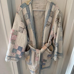 May include: A quilted robe with a patchwork design in shades of white, blue, pink, and yellow. The robe has a tie belt, pockets, and is hanging on a white hanger against a white door.