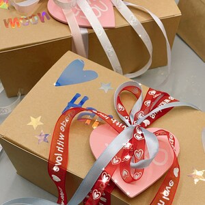 May include: Two brown gift boxes with decorative ribbon and hearts. The top box has a white ribbon with the word "Love" on it. The bottom box has a red ribbon with the words "Love with love" on it. Both boxes have a pink heart with the word "Love" on it.