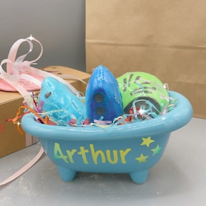 May include: A light blue ceramic bathtub filled with colourful bath bombs and shredded paper. The tub has the name "Arthur" written on the side. A brown paper bag with a logo is in the background.