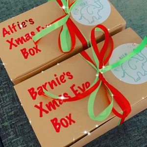 May include: Two rectangular gift boxes with red text reading "Alfie's Xmas Box" and "Barnie's Xmas Eve Box". Each box is tied with red and green ribbons and has a circular sticker with an elephant logo.