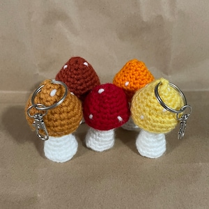 May include: Five crocheted mushroom keychains in different colors. The mushrooms are red, orange, yellow, and brown with white spots. The keychains are attached to silver key rings.