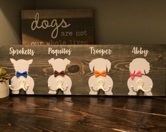 May include: A wooden sign with four dog silhouettes, each with a different colored bow tie. The sign has the names "Sproketts", "Paquitos", "Trooper", and "Abby" written on it. The sign has two hooks on each silhouette, making it a dog leash holder.