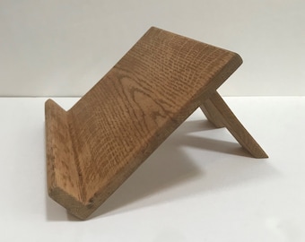 Puede incluir: Un soporte de libro de madera de roble marrón claro con un diseño sencillo. El soporte está inclinado para sostener un libro o una tableta.