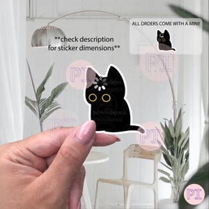 May include: A black cat sticker with yellow eyes and a white flower detail on its head is held up. The sticker is in front of a window with text that reads "ALL ORDERS COME WITH A MINI!" and "check description for sticker dimensions**".