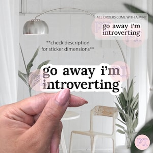 以下が含まれることがあります： 太字の黒いフォントで「go away i'm introverting」というテキストが入った白いステッカー。ステッカーは手で持たれています。追加のテキストには「check description for sticker dimensions」と「ALL ORDERS COME WITH A MINI!」が含まれています。