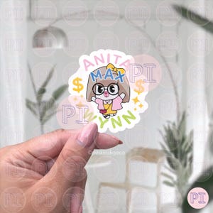 Op de afbeelding: Een sticker met een stripfiguur met een bril, een gele jurk en een roze jasje. De sticker heeft de tekst "ANITA MAX WYNN" in verschillende kleuren, met dollartekens en sterren. De sticker wordt door een hand vastgehouden.