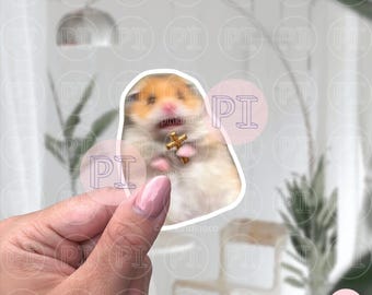 May include: A sticker of a hamster holding a small gold cross. The hamster is looking up with a surprised expression. The sticker is on a white background with a hand holding it.