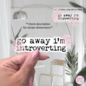 以下が含まれることがあります： 「go away i'm introverting」のテキストが黒の擦り切れたフォントで書かれた白いステッカー。ステッカーは人の手で持たれています。追加のテキストには「check description for sticker dimensions」と「ALL ORDERS COME WITH A MINI!」が含まれています。