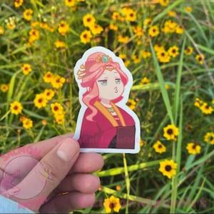 May include: A sticker featuring a cartoon character with pink hair and a red and gold outfit. The character has a crown and is holding a book. The sticker has the text "MoonMoonArt" on it.