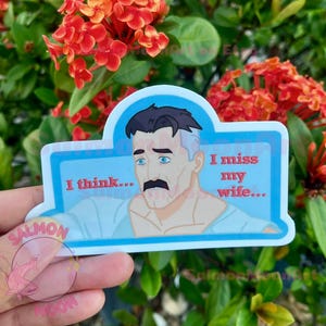May include: A sticker featuring a cartoon character with a mustache and the text "I miss my wife...". The sticker has a blue border and is held in front of a background of red flowers and green leaves.