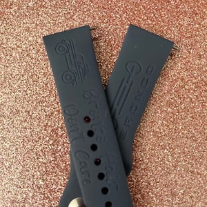 Puede incluir: Dos correas de reloj de silicona gris oscuro con un dise&ntilde;o de coche vintage y el texto "Well, Well, Well" y "The Big NC" en relieve.