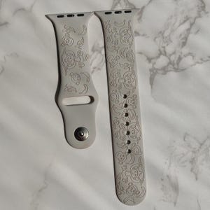Puede incluir: Una correa de reloj de silicona gris claro con un patrón en relieve de monos y plátanos. La correa de dos piezas incluye un cierre de pasador metálico en un lado y una serie de orificios de ajuste en el otro para un reloj inteligente.