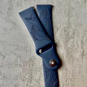K&ouml;nnte beinhalten: Ein dunkelblaues Silikon-Armband mit einem Wal-Design. Das Armband hat eine silberne Schnalle.