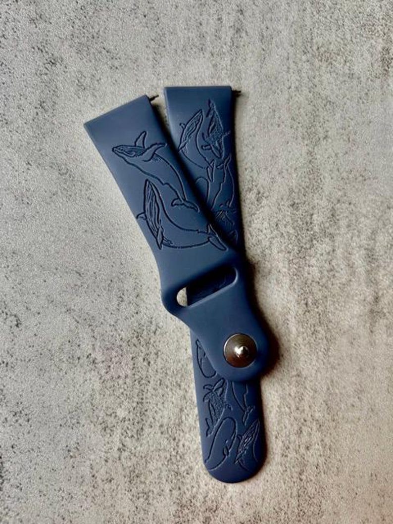 K&ouml;nnte beinhalten: Ein dunkelblaues Silikon-Armband mit einem Wal-Design. Das Armband hat eine silberne Schnalle.