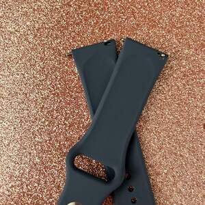 Puede incluir: Dos correas de reloj de silicona gris oscuro con una hebilla plateada. Las correas est&aacute;n cruzadas una sobre la otra.