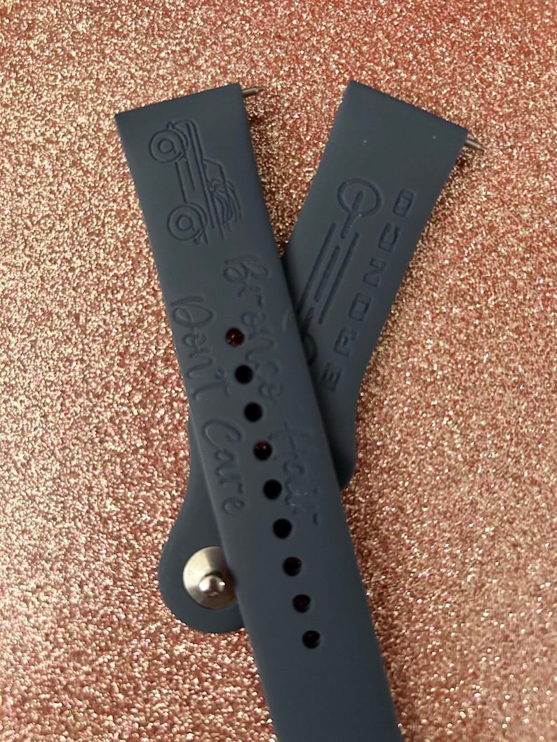 Puede incluir: Dos correas de reloj de silicona gris oscuro con un dise&ntilde;o de coche vintage y el texto "Well, Well, Well" y "The Big NC" en relieve.