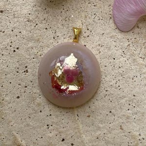 Op de afbeelding: Een ronde hanger met een goudkleurige beugel. De hanger is lichtroze met bladgoud en een roze bloemenontwerp. De hanger ligt op een getextureerd, lichtgekleurd oppervlak.