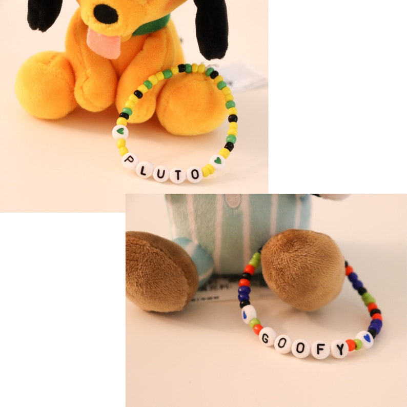 以下が含まれることがあります： ビーズのブレスレットが付いたぬいぐるみ犬が2匹。黄色の犬は、緑、黄、黒のビーズのブレスレットに「PLUTO」と書いてあります。青と白のストライプの犬は、赤、オレンジ、青、黒のビーズのブレスレットに「GOOFY」と書いてあります。