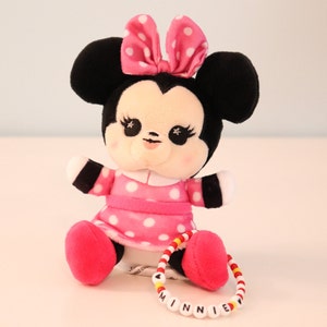 以下が含まれることがあります： ピンクと黒のぬいぐるみミニーマウスのドールで、ピンクのドット柄のドレスとピンクのリボンをしています。ドールは白い表面に座っており、赤いビーズと黄色のビーズでできたブレスレットをしています。ブレスレットには黒い文字で「MINNIE」と書いてあります。