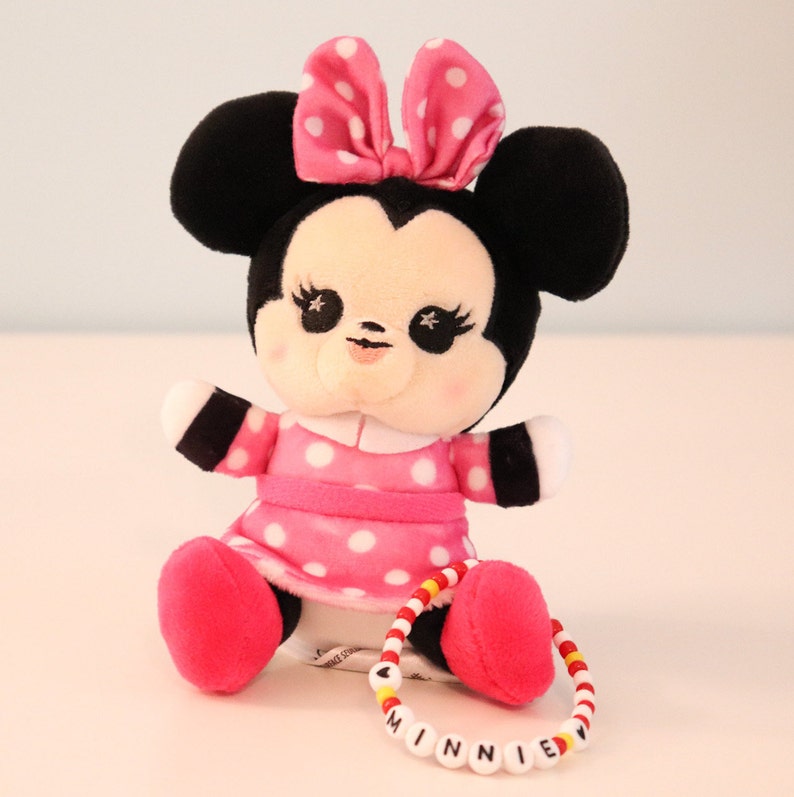 以下が含まれることがあります： ピンクと黒のぬいぐるみミニーマウスのドールで、ピンクのドット柄のドレスとピンクのリボンをしています。ドールは白い表面に座っており、赤いビーズと黄色のビーズでできたブレスレットをしています。ブレスレットには黒い文字で「MINNIE」と書いてあります。