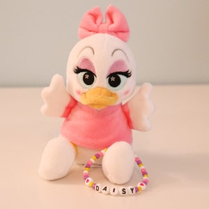 以下が含まれることがあります： ピンクの服を着てピンクのリボンをつけた白いぬいぐるみのかわいいアヒル。アヒルは刺繍されたまつげと黄色いくちばしをしています。アヒルは「DAISY」と書かれたビーズのブレスレットをしています。