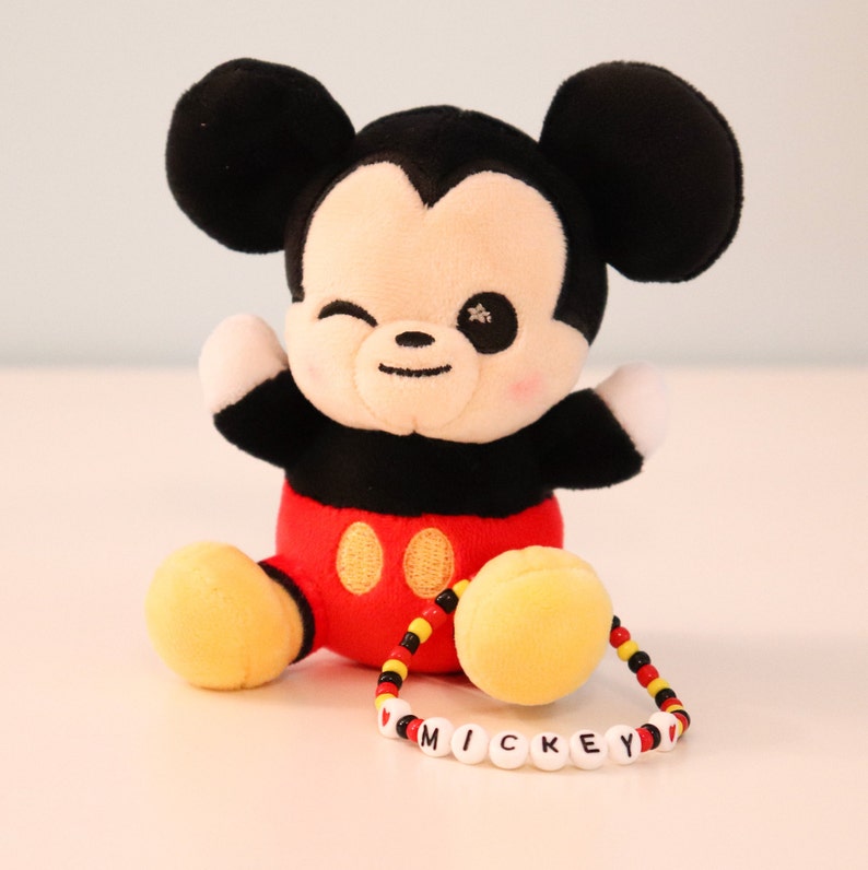 以下が含まれることがあります： 赤い服を着たミッキーマウスのぬいぐるみ。黄色いビーズのブレスレットには、白い文字で「I love Mickey」と書かれています。