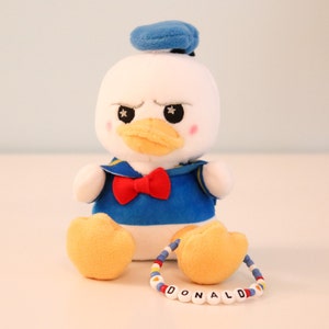 以下が含まれることがあります： 青いセーラーシャツに赤い蝶ネクタイ、青いセーラーハットを被ったドナルドダックのぬいぐるみ。おもちゃは白い体で、黄色い足と、白い文字で「DONALD」と綴られたビーズのブレスレットをしています。