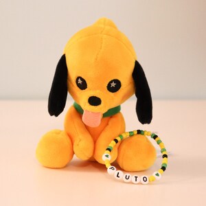 以下が含まれることがあります： 黒い耳と緑色の首輪が付いた黄色のぬいぐるみ犬の玩具。犬はピンクの舌を出しています。玩具は、「PLUTO」という文字が白いビーズで書かれたビーズブレスレットの隣に置かれています。