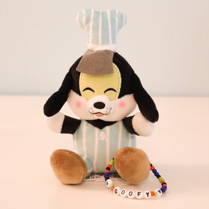 以下が含まれることがあります： 青と白のストライプのシェフハットとパジャマを着た、プルートのぬいぐるみ。おもちゃは足を組んで座っており、足首に「GOOFY」と書かれたビーズのブレスレットをしています。