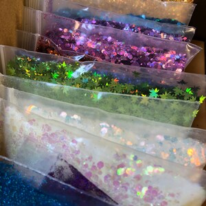 K&ouml;nnte beinhalten: Eine Sammlung von buntem Glitter in kleinen Plastikt&uuml;ten. Der Glitter ist in einem Regenbogenmuster angeordnet, mit gr&uuml;nem, goldenem, violettem, gr&uuml;nem, wei&szlig;em, blauem und orangefarbenem Glitter.
