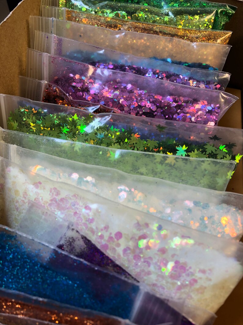 K&ouml;nnte beinhalten: Eine Sammlung von buntem Glitter in kleinen Plastikt&uuml;ten. Der Glitter ist in einem Regenbogenmuster angeordnet, mit gr&uuml;nem, goldenem, violettem, gr&uuml;nem, wei&szlig;em, blauem und orangefarbenem Glitter.