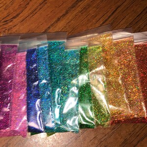 K&ouml;nnte beinhalten: Sortiment an schimmerndem Glitter in verschiedenen Farben, verpackt in durchsichtigen Plastikt&uuml;ten. Der Glitter ist in einem Regenbogenmuster angeordnet, mit Rot, Orange, Gelb, Gr&uuml;n, Blau und Lila.