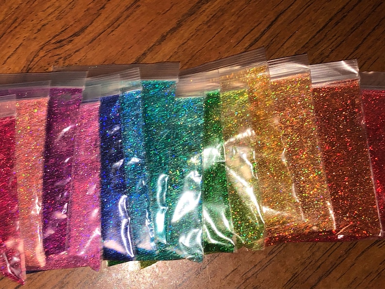 K&ouml;nnte beinhalten: Sortiment an schimmerndem Glitter in verschiedenen Farben, verpackt in durchsichtigen Plastikt&uuml;ten. Der Glitter ist in einem Regenbogenmuster angeordnet, mit Rot, Orange, Gelb, Gr&uuml;n, Blau und Lila.